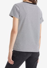 Kobieta w krótkim rękawie, w czarno-białą poziomą w paski koszulce i czarnych dżinsach, pokazana od tyłu na białym tle.
