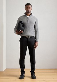 Suéter gris acanalado con un cuello alto, pantalones negros y zapatos gruesos negros. El modelo sostiene un bolso negro, de pie sobre un suelo de madera.