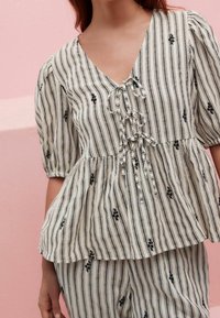 Blouse peplum en tissu léger à rayures, avec un décolleté en V profond, des manches bouffantes et une broderie florale noire sur fond crème.