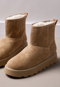 Beżowe zamszowe botki za kostkę z miękkim wnętrzem, ozdobnymi przeszyciami wzdłuż szwów oraz grubą, teksturowaną podeszwą zapewniającą stabilność.