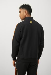 Sweat-shirt noir avec un col haut, comportant des rayures orange et jaunes le long des manches. Petit label "NTS" à l'arrière du cou. Texture douce.