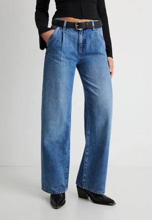 Oseba v širokih modrih kavbojkah z visokim pasom in črnim pasom, črni krajši majici ter črnih škornjih z visokim petnim in zoženim koncem, z rokami v žepih.