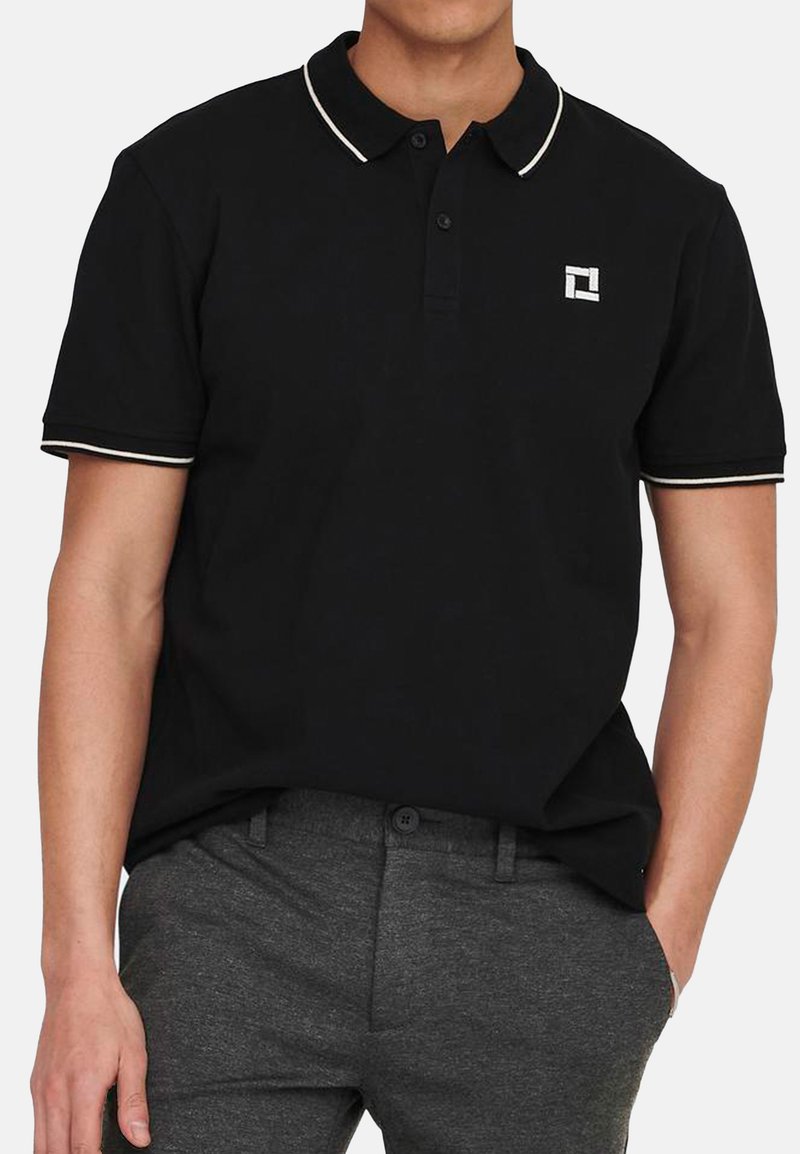 Schwarzes Poloshirt aus Baumwolle, mit einem weißen Kragen und Ärmelabschlüssen, sowie einem Logo im oberen linken Bereich. Kurze Ärmel und eine Knopfleiste.