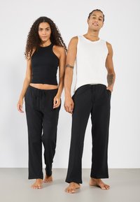 Débardeur noir et pantalon à cordon pour elle, chemise sans manches blanche et pantalon noir pour lui, le tout en tissu doux, présenté sur un fond neutre.