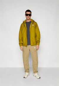 Kollane veekindel jakk kapuutsiga, hall T-särk, beežid püksid ja heledad tossud. Märgatav logo jakil ja pükstel.