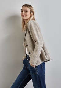 Texturierte beige Blazer mit zwei schwarzen Knöpfen, der eine taillierte Passform und eine offene Front hat, kombiniert mit dunkelblauen Jeans. Neutrale Kulisse.
