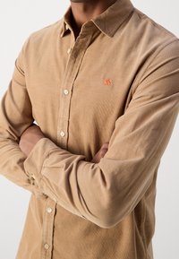 Hellbraunes Cordhemd mit Knopfkragen, Frontknopfleiste und einem orangefarbenen gestickten Logo auf der Brust.