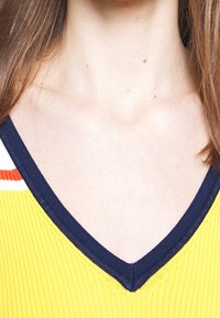 Maglione giallo a coste con scollatura a V blu navy e strisce arancioni e bianche lungo il colletto. Inquadratura ravvicinata della scollatura e delle spalle.