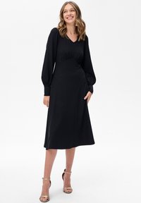 Kobieta uśmiechająca się, ubrana w czarną midi sukienkę z długimi rękawami, z marszczoną talią i detalami przy mankietach, w połączeniu z beżowymi szpilkami na kostkę.