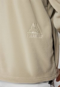 Jachetă din fleece Polartec bej, cu o textură moale, având un logo brodat, buzunare laterale cu fermoar și o margine ușor curbată.