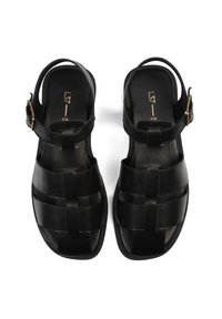 Sandalias de cuero negro con un diseño trenzado, punta abierta, correa ajustable en el tobillo y una textura suave. Detalle de hebilla con acentos dorados.