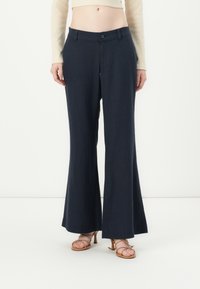 Pantaloni a gamba larga blu navy realizzati in tessuto leggero, con chiusura a bottone e tasche frontali. Indossati con sandali marrone chiaro a strap.