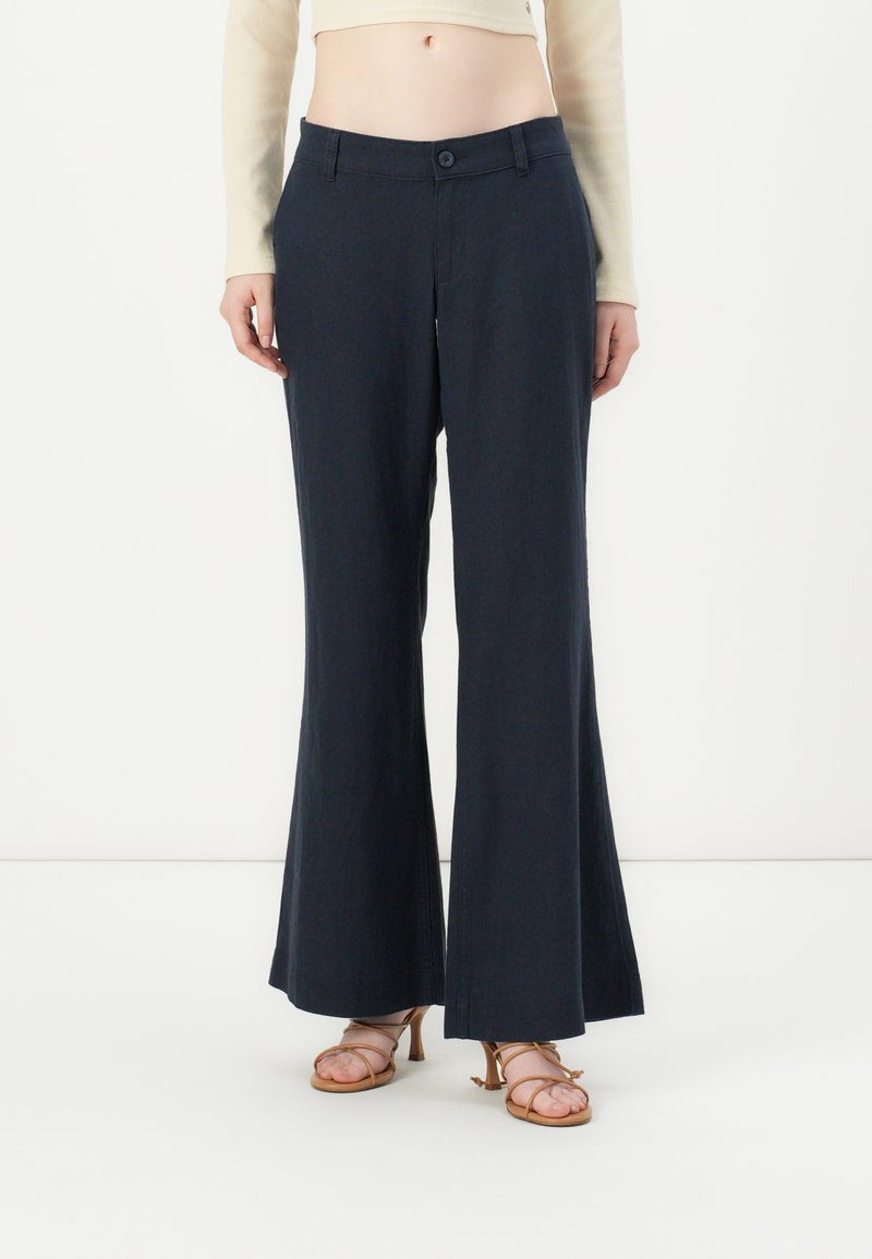 Pantaloni a gamba larga blu navy realizzati in tessuto leggero, con chiusura a bottone e tasche frontali. Indossati con sandali marrone chiaro a strap.