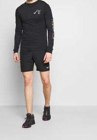 Schwarzes Langarm-Nike-Shirt mit Grafik und farbigem Text auf den Ärmeln, kombiniert mit schwarzen Shorts und schwarzen Sneakers mit lila Akzenten.