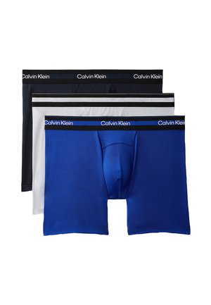 Skrętny mężczyzna stoi boso, ubrany w czarne bokserki Calvin Klein na jednolitym szarym tle.