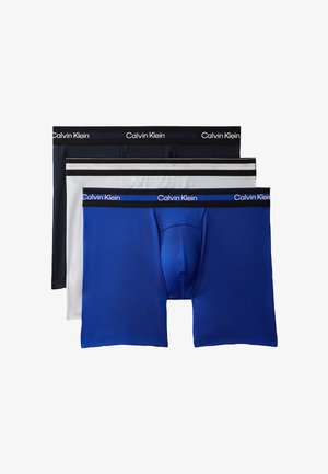 Skrętny mężczyzna stoi boso, ubrany w czarne bokserki Calvin Klein na jednolitym szarym tle.