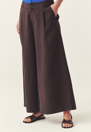 Žena v tmavohnedých širokých nohaviciach s prednými záhybmi a vreckami, v kombinácii s čiernymi plochými sandálmi, stojaca pred jednoduchým pozadím.