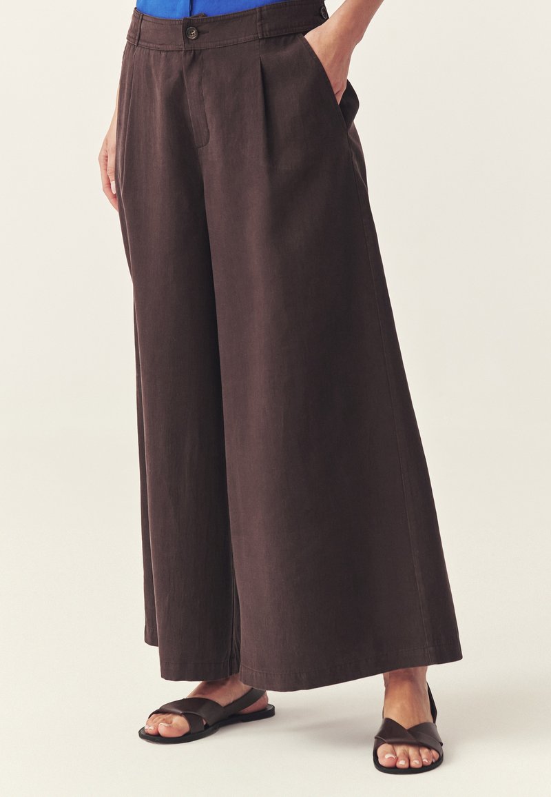 Femme portant un pantalon large marron foncé avec des plis devant et des poches, associé à des sandales plates noires, debout devant un fond uni.