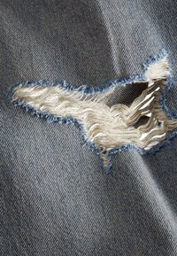 Il tessuto di denim blu strappato mette in evidenza un strappo con bordi sfrangiati e fili bianchi. La texture è morbida, con un classico motivo intrecciato.