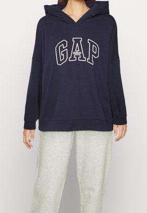 Person iført en mørkeblå hoodie med teksten "GAP Athletic" og lysegrå sweatpants, stående mod en ensfarvet baggrund.