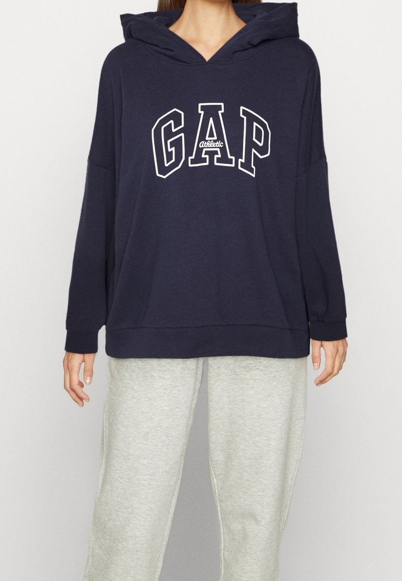 Personne portant un sweat à capuche bleu marine avec le texte "GAP Athletic" et un pantalon de survêtement gris clair, debout devant un fond uni.