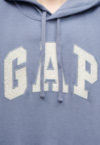 Κοντινό πλάνο ενός ανοιχτό μωβ φούτερ με κουκούλα, με λευκά ανάγλυφα γράμματα "GAP" στο στήθος και κορδόνια στην κουκούλα που ταιριάζουν στο χρώμα.
