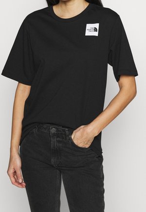 Kvinde iført en sort North Face t-shirt med korte ærmer og sorte jeans, den ene hånd i lommen, mod en ensfarvet lys baggrund.