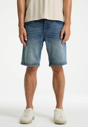 Mann trägt ausgewaschene blaue Jeansshorts und weiße Turnschuhe, steht vor einem schlichten weißen Hintergrund.