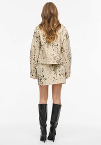 Veste en denim beige avec motif éclaboussé noir, associée à une jupe assortie. Le mannequin porte des bottes noires montant jusqu'aux genoux. Tissu texturé, coupe décontractée.