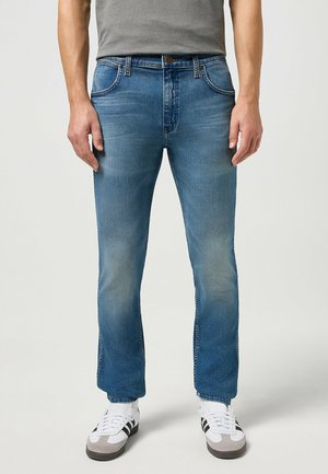 Jeans de denim azul claro con corte recto, un sutil desgastado, cinco bolsillos y herrajes en bronce, combinados con zapatillas blancas.