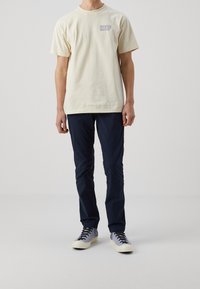 Krémové bavlněné tričko s modrým logem "HUF", doplněné o tmavě modré úzké kalhoty a šedé tenisky s béžovými podrážkami a černými tkaničkami.