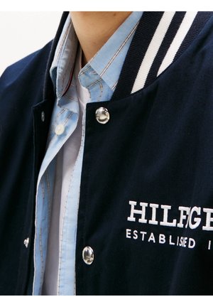 Personne portant une veste universitaire bleu marine avec le texte "HILFIGER ESTABLISHED" sur une chemise bleu clair partiellement déboutonnée.