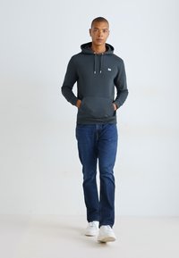 Donkergrijze hoodie met een kangoeroezak en verstelbare trekkoorden, gecombineerd met blauwe jeans en witte sneakers, tegen een lichte achtergrond.