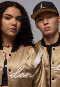 Giacche in satin dorato con accenti neri, dotate di ricami con il testo "Jordan" e dettagli bianchi, indossate da due persone con accessori complementari.