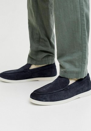 Hombre con zapatos tipo slip-on de ante azul oscuro con suelas blancas y calcetines beige, combinados con pantalones casuales de color verde oliva.