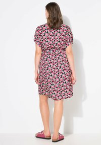 Abito nero con maniche corte, caratterizzato da un motivo a cuori rosa e verdi. Dettaglio con cintura in vita e design fino al ginocchio. Indossato con sandali rosa.