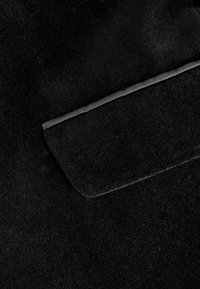 Tela de ante negro con una textura suave, que presenta un pequeño bolsillo bordeado con un fino ribete negro texturizado.
