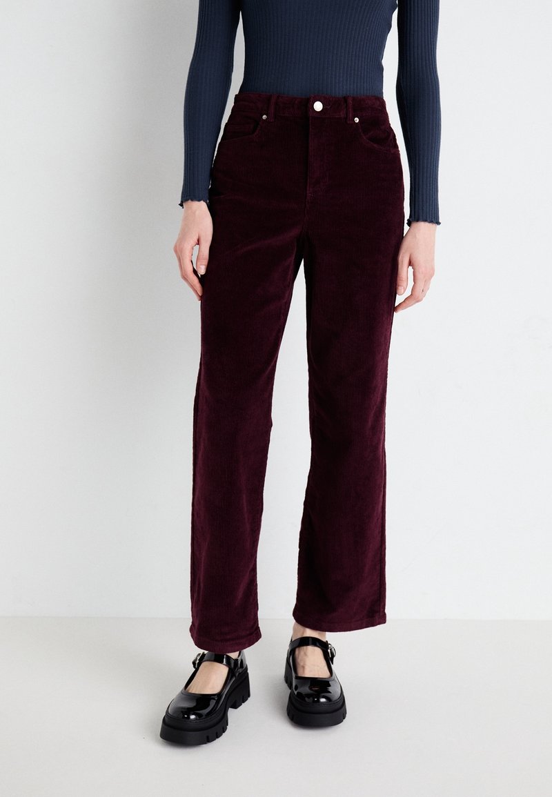 Pantalon en velours côtelé bordeaux avec une silhouette à jambes droites, doté de poches avant et de boutons en argent, porté avec des chaussures à plateforme noires.