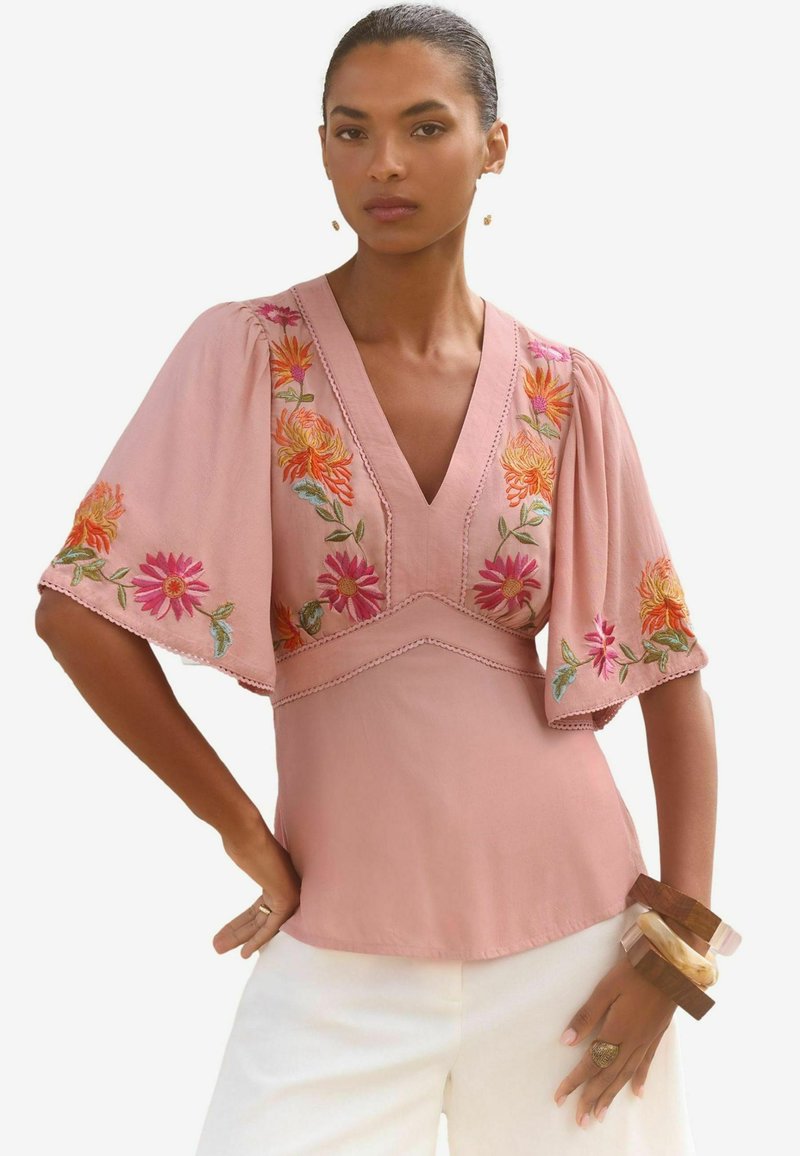 Blusa rosa con scollatura a V e maniche corte e svasate. Presenta ricami floreali colorati lungo le maniche e il corpetto. Tessuto leggero.