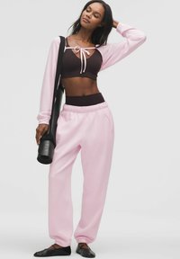 Világos rózsaszín, hosszú ujjú felső, ami megkötős elülső részes a sötétbarna sportmelltartó felett; hozzáillő rózsaszín tréningnadrág; fekete balettcipők; egy fekete jógamatracot tart.