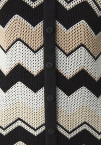 Cardigan en maille avec un motif en chevron noir, beige et blanc. Il présente une finition texturée et des boutons noirs sur le devant.