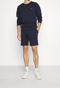 Homme portant un sweat-shirt bleu marine et un short assorti, avec des chaussettes blanches et des baskets blanches, se tenant devant un fond blanc uni.