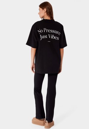 Kobieta zwrócona tyłem, ubrana w czarną oversize koszulkę z napisem "No Pressure Just Vibes" oraz czarne spodnie i beżowe buty na platformie.