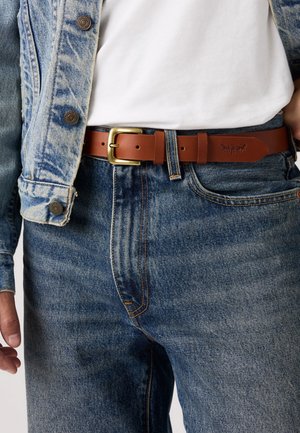 Hnědý kožený opasek se zlatou kovovou sponou, nošený přes modré džínové kalhoty. Opasek má hladkou strukturu a zaoblené konce.