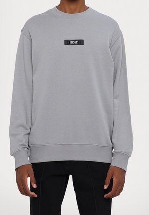 Sudadera gris con mangas largas, cuello redondo y puños acanalados. Presenta un parche de logo rectangular negro en el pecho. Textura suave.