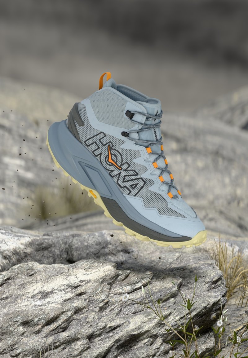 Turistická bota Hoka se světlým šedým syntetickým svrškem, síťovanými detaily, oranžovými tkaničkami a texturovanou podrážkou v šedé a žluté barvě.