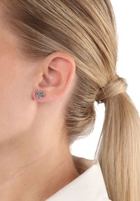 Närbild av en kvinnas öra med blont hår uppsatt, som bär en liten silverfärgad rosformad studörhänge.