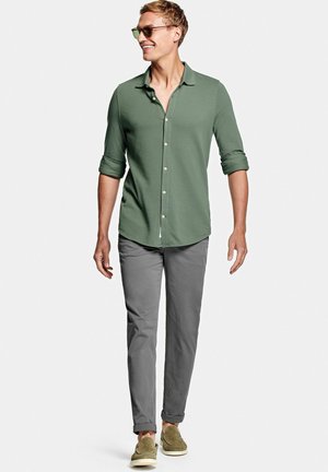 Uomo con camicia verde abbottonata, pantaloni grigi, occhiali da sole e scarpe verdi che cammina e sorride su uno sfondo chiaro e semplice.