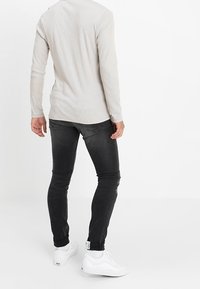 Camisa de manga larga gris claro, diseño ajustado, combinada con jeans ajustados negros y zapatillas blancas. Detalles notables: tejido texturizado y etiqueta del logo en la parte trasera.
