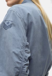 Κοντινό πλάνο ενός ατόμου που φοράει μπλε μπουφάν με κεντημένο αστέρι και έμβλημα σε σχήμα βέλους στον ώμο, με μακριά ξανθά μαλλιά ορατά.